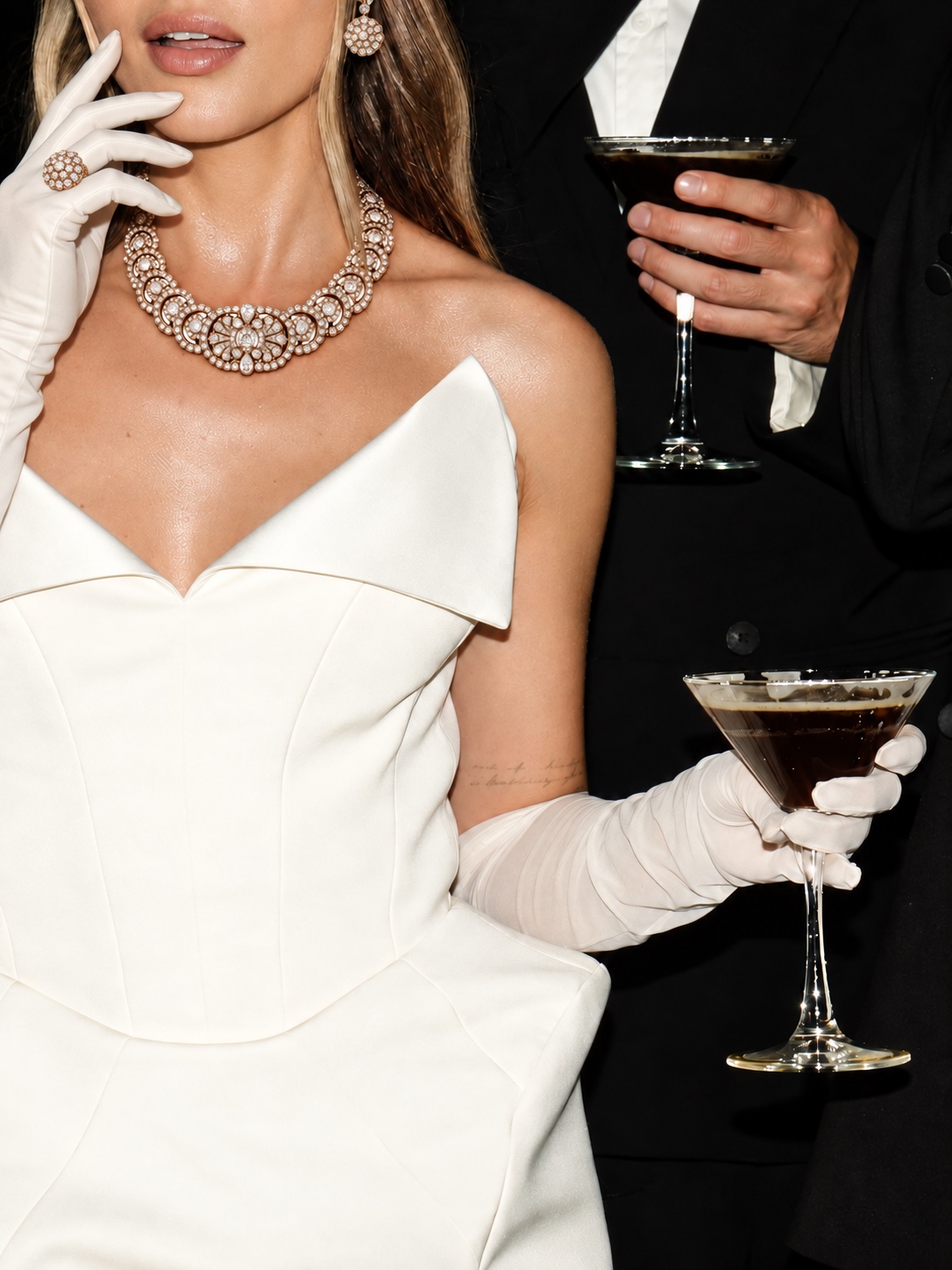 a close-up of a woman in an ivory sculptural gown and gloves, wearing floral diamond jewellery and holding a cocktail, beside a man in black evening wear.